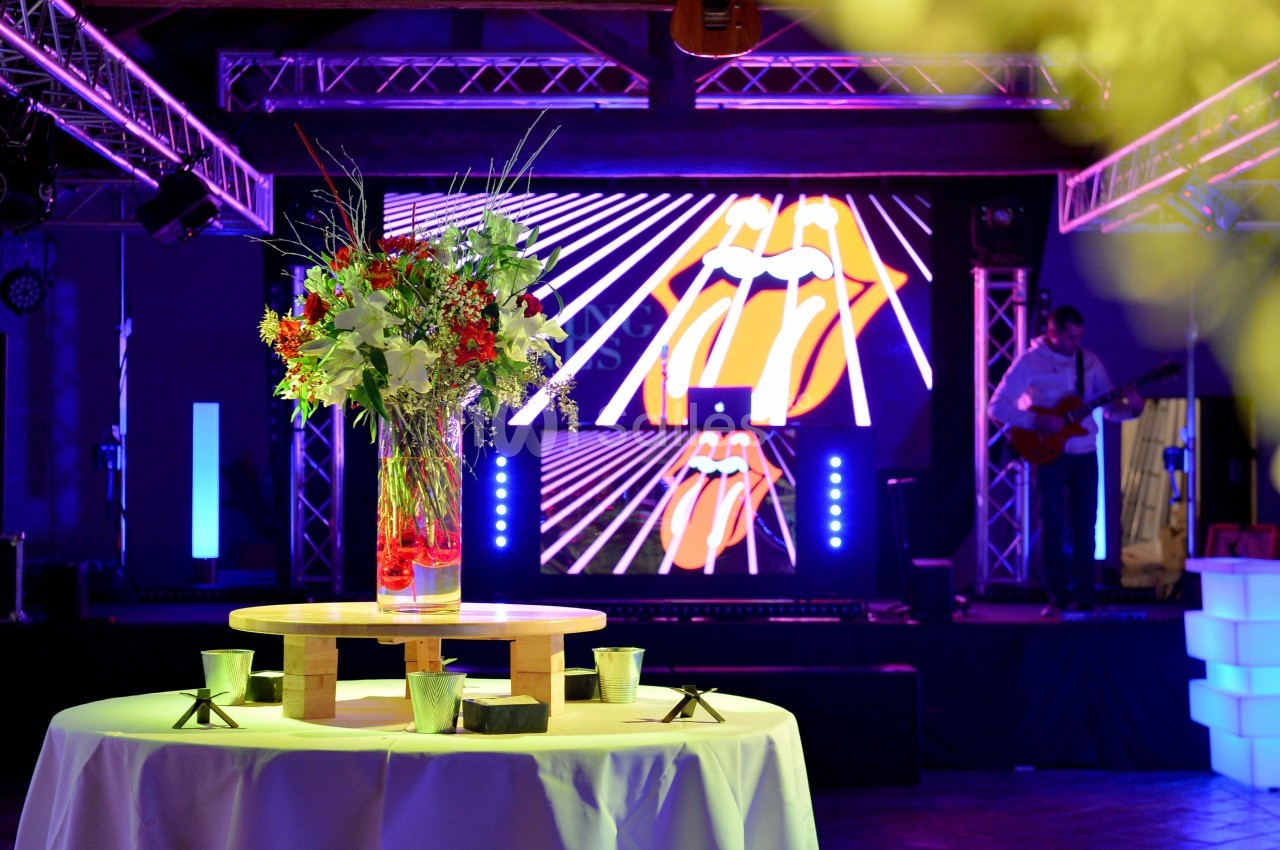 Vase de fleurs sur une table ronde devant une scène éclairée avec écran lumineux et équipement de DJ.