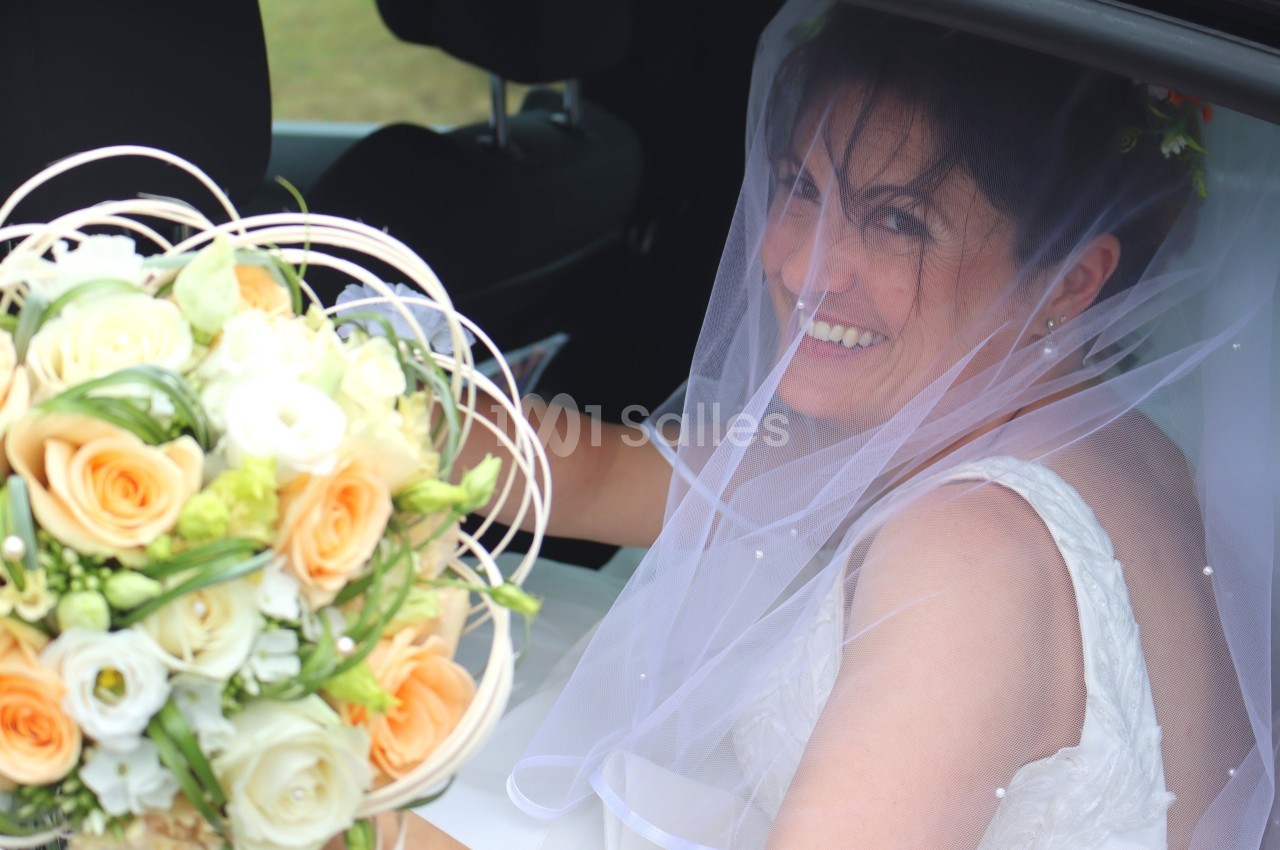 Une mariée souriante en robe blanche et voile, tenant un bouquet de fleurs pastel, assise dans une voiture.