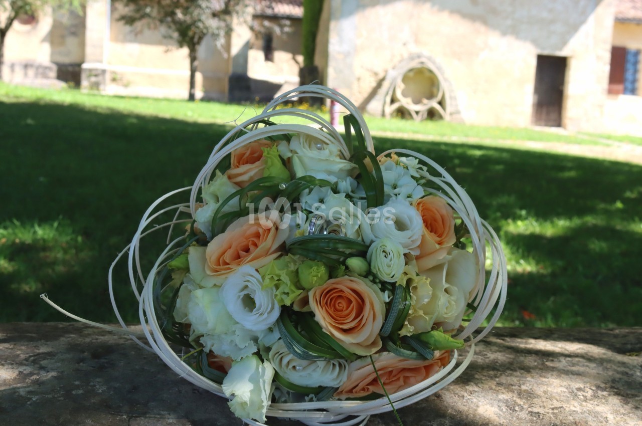 Bouquet de fleurs aux tons orange et blanc posé sur une pierre, avec un jardin et un bâtiment en arrière-plan.