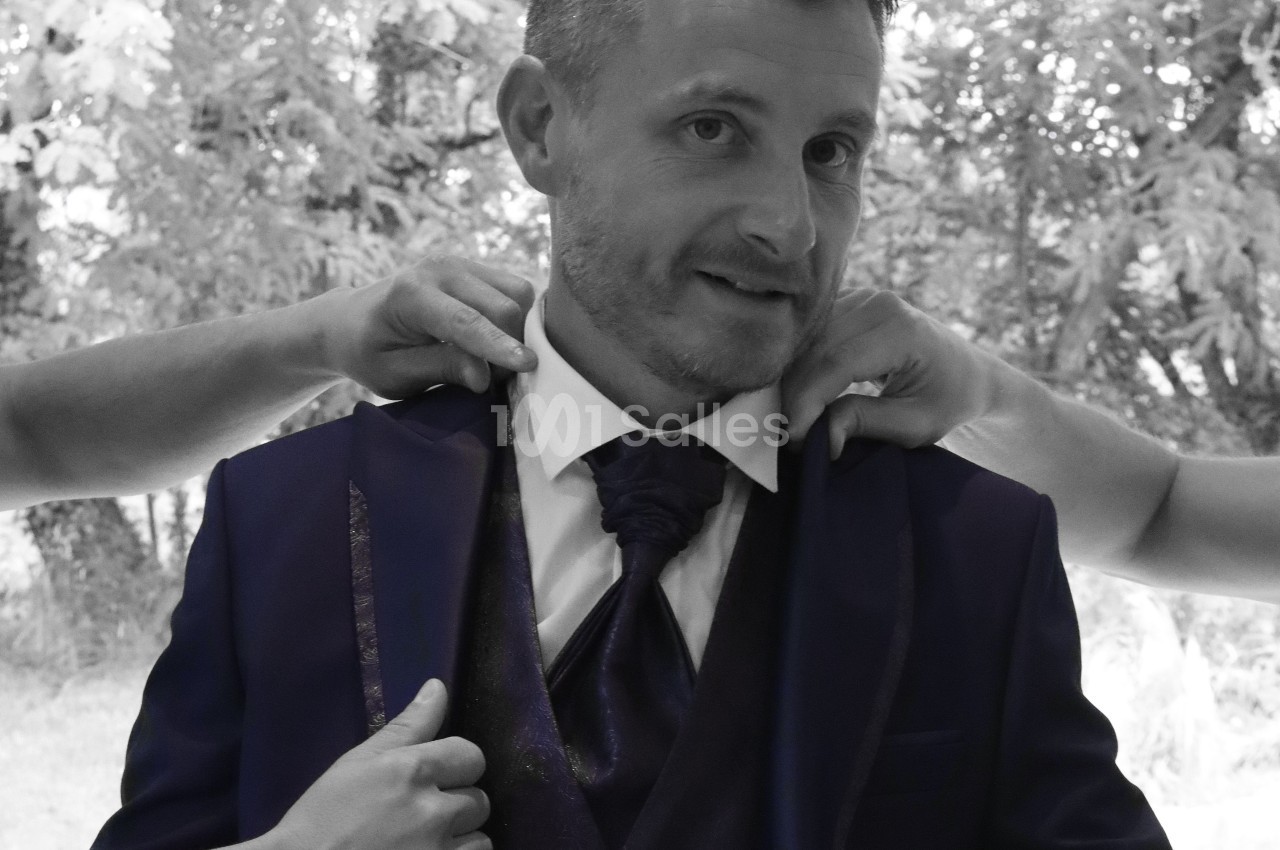 Un homme en costume sombre est aidé par deux personnes ajustant sa veste, devant un fond naturel flou.