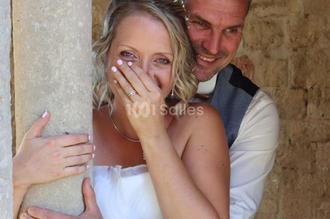 Un couple souriant en tenue de mariage, la mariée couvrant partiellement son visage avec sa main.