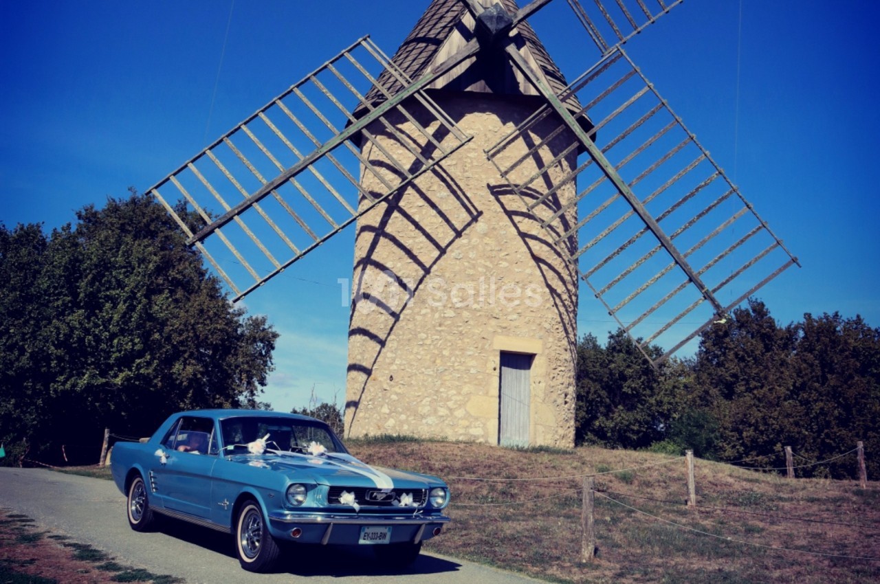 Voiture ancienne bleue garée devant un moulin en pierre avec des ailes en bois, entouré de végétation.