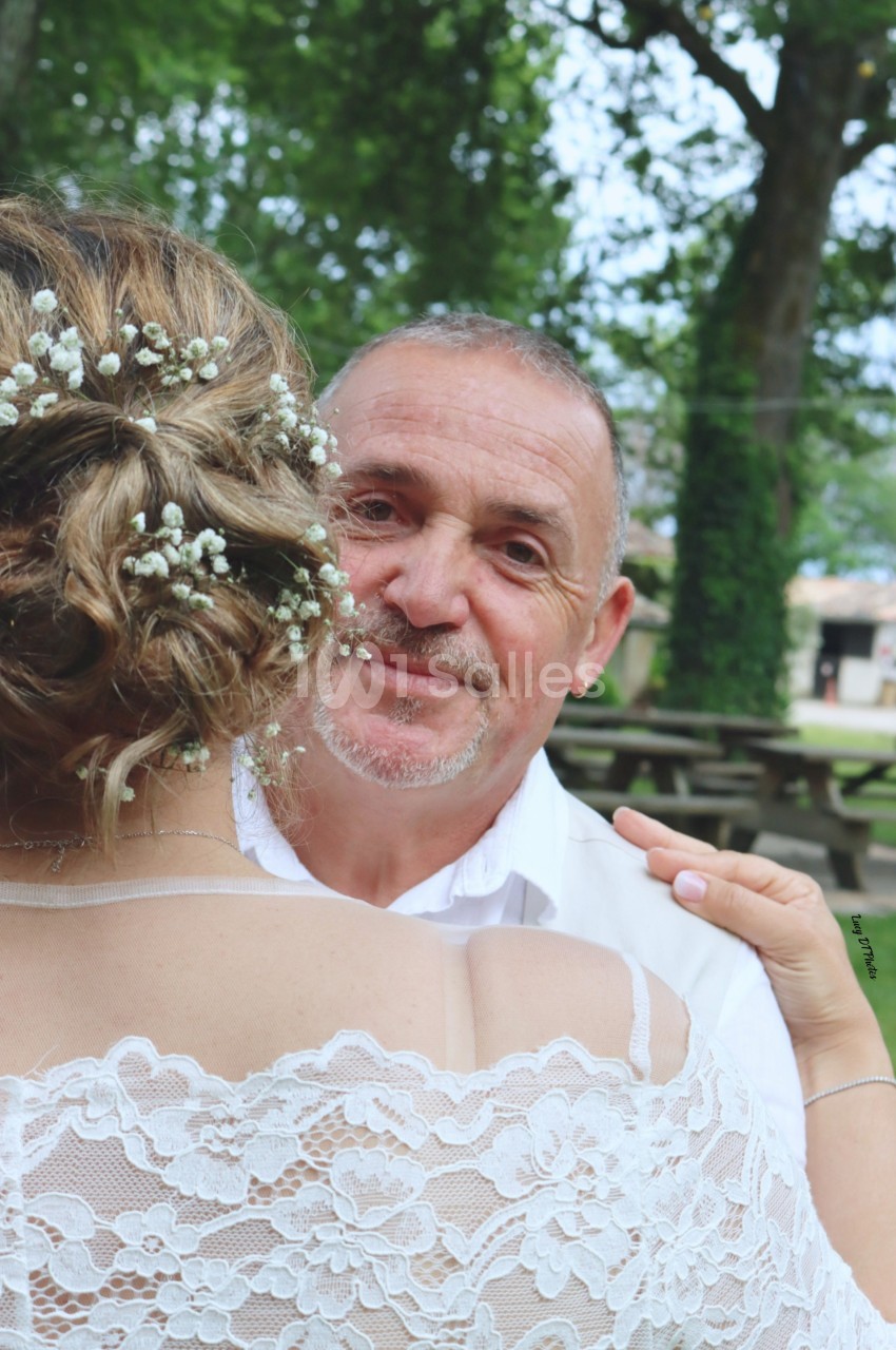 Un homme souriant regarde vers l'objectif, tenant une femme en robe de dentelle blanche dans un parc verdoyant.