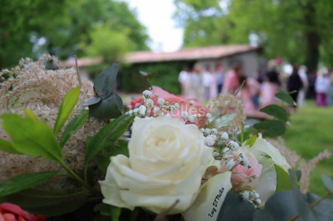 Bouquet de fleurs blanches et roses au premier plan, avec des personnes floues en arrière-plan dans un jardin verdoyant.