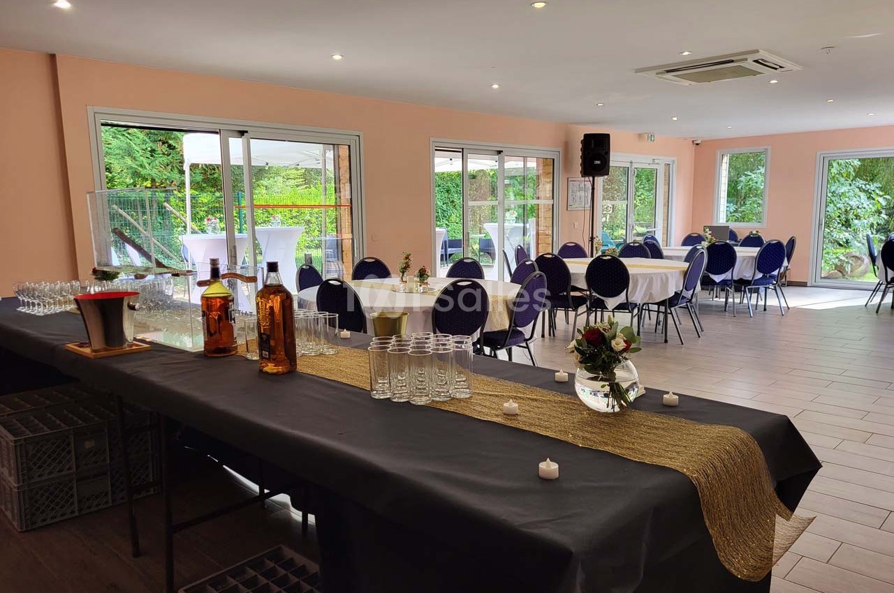 Salle lumineuse avec tables rondes dressées, chaises bleues et buffet décoré, vue sur un jardin à travers de grandes baies…