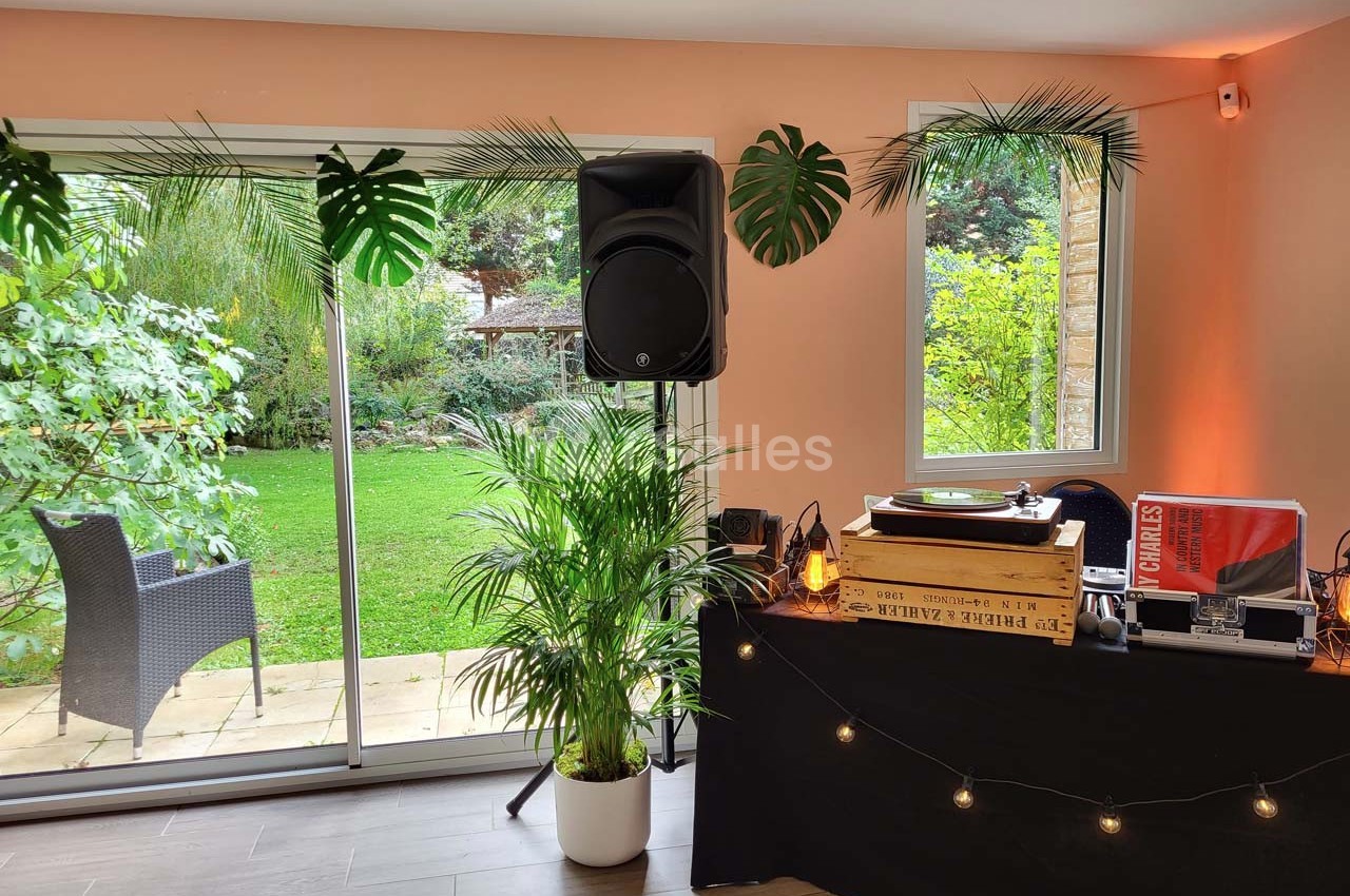 Table de DJ avec platine vinyle et enceintes près d'une baie vitrée donnant sur un jardin verdoyant.