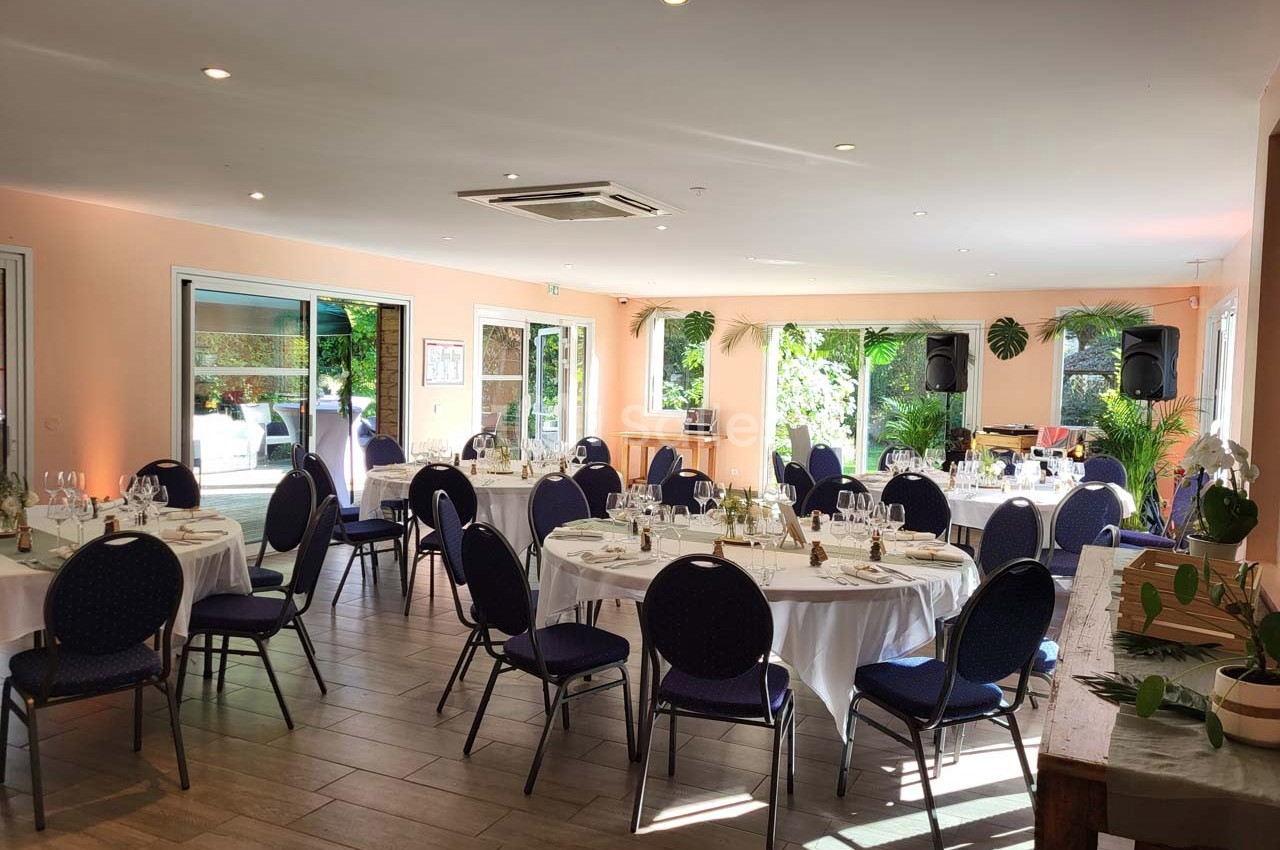 Salle lumineuse aménagée pour un événement, avec des tables rondes dressées et des chaises noires, vue sur un jardin.