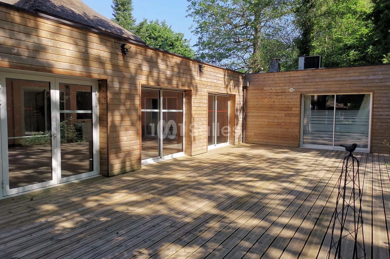 Terrasse en bois entourée d'une maison en bardage bois avec de grandes baies vitrées, sous un ciel dégagé.