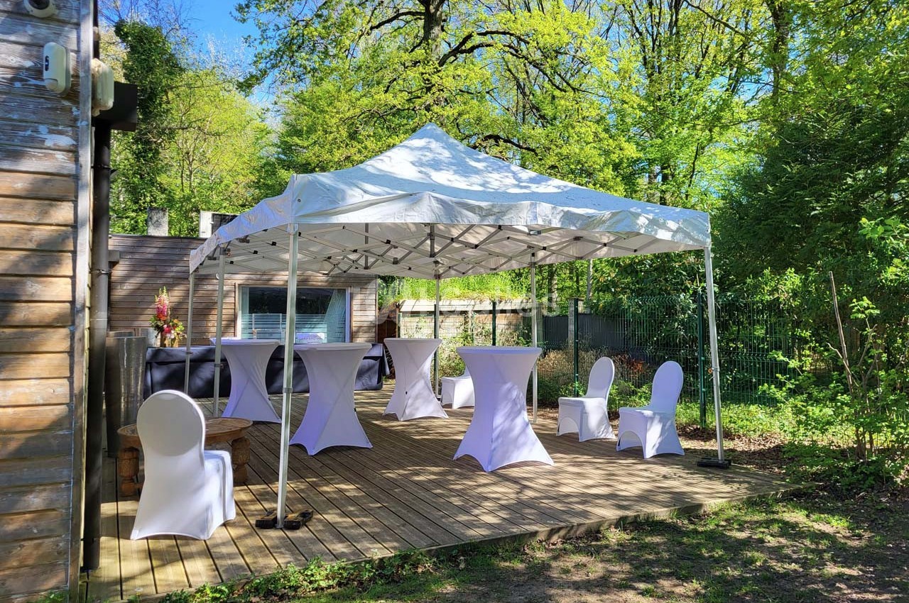 Tente blanche installée sur une terrasse en bois, entourée de verdure, avec tables et chaises habillées de housses.