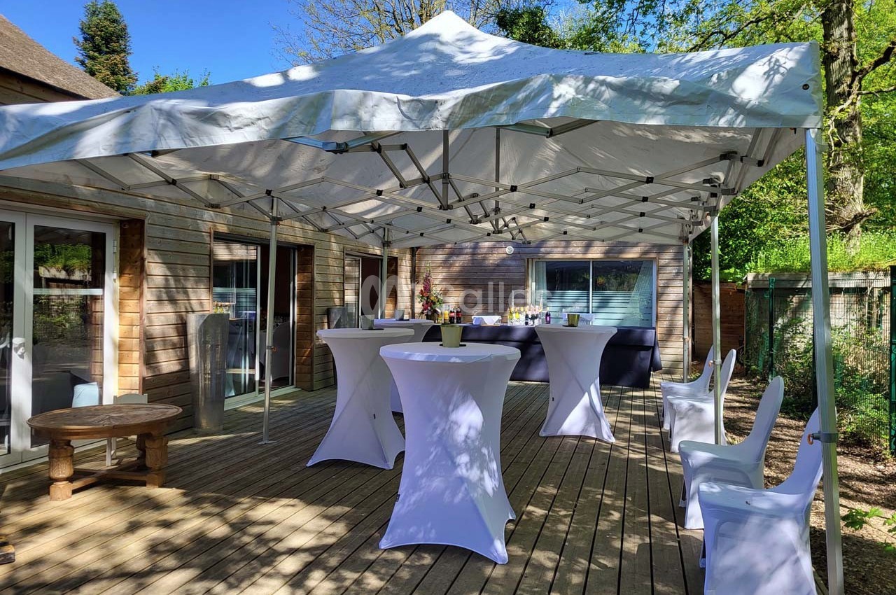 Tonnelle blanche installée sur une terrasse en bois, avec tables hautes et chaises, entourée de verdure.