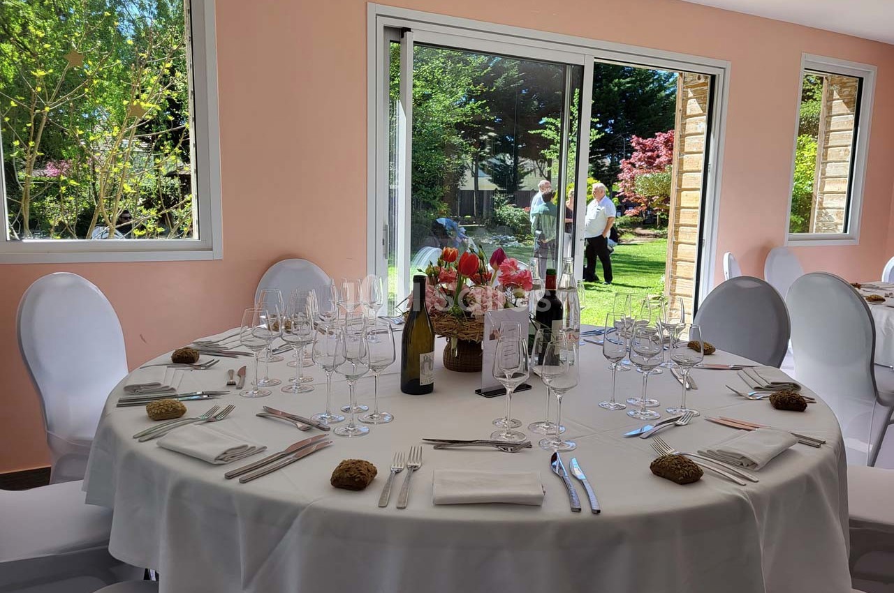 Table ronde dressée avec nappes blanches, verres et couverts, près de baies vitrées donnant sur un jardin verdoyant.