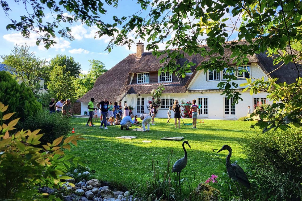 Un groupe de personnes joue et discute dans le jardin d'une maison à toit de chaume, entourée de verdure.