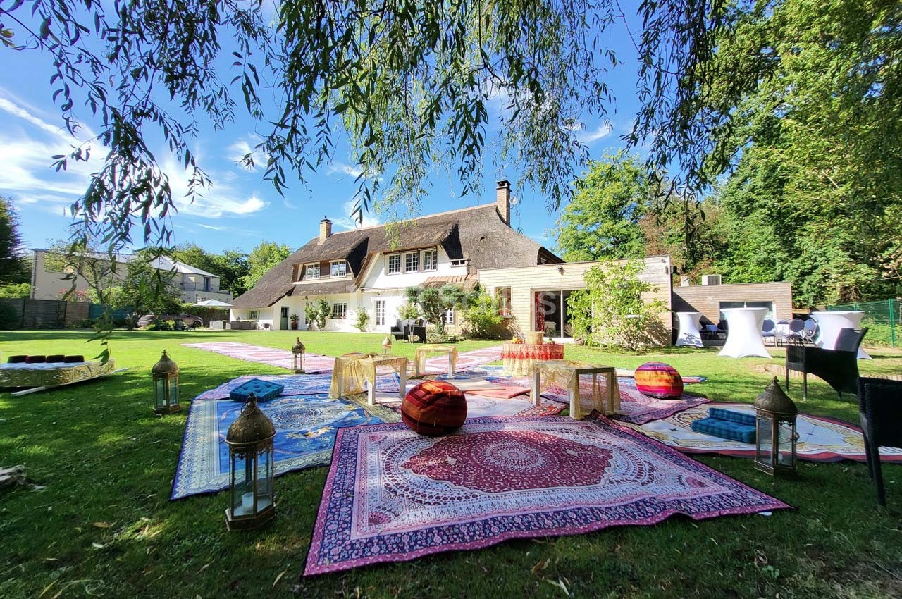 Jardin avec tapis colorés, coussins et lanternes devant une maison à toit de chaume entourée de verdure.