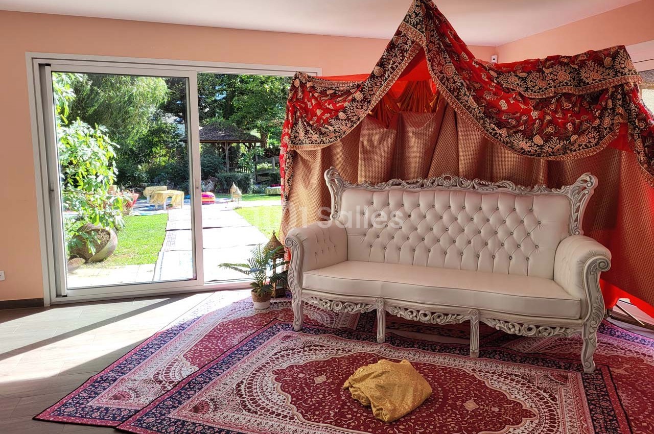 Canapé blanc devant une tente décorative rouge et dorée, posé sur des tapis orientaux, avec vue sur un jardin ensoleillé.