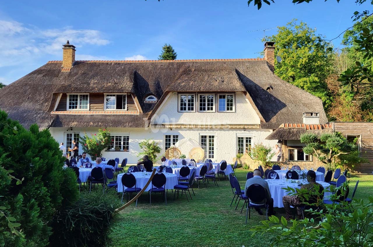 Tables dressées dans un jardin verdoyant devant une maison à toit de chaume, sous un ciel bleu.