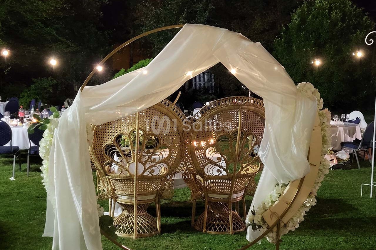 Deux fauteuils en rotin ornés de fleurs et drapés blancs, disposés dans un cercle métallique sur une pelouse en soirée.