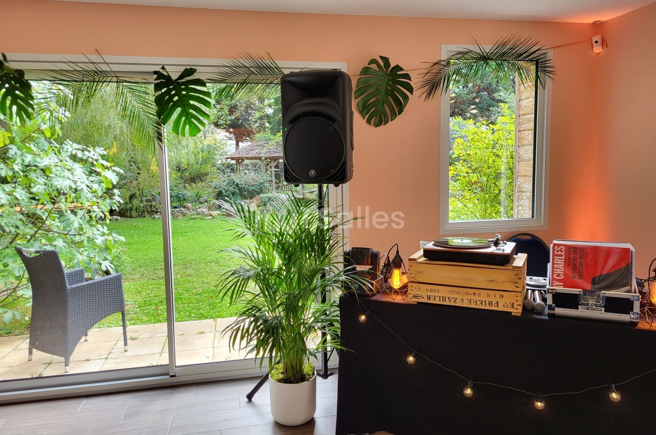 Table de mixage avec platine vinyle et enceinte dans une pièce lumineuse, vue sur un jardin verdoyant par une baie vitrée.