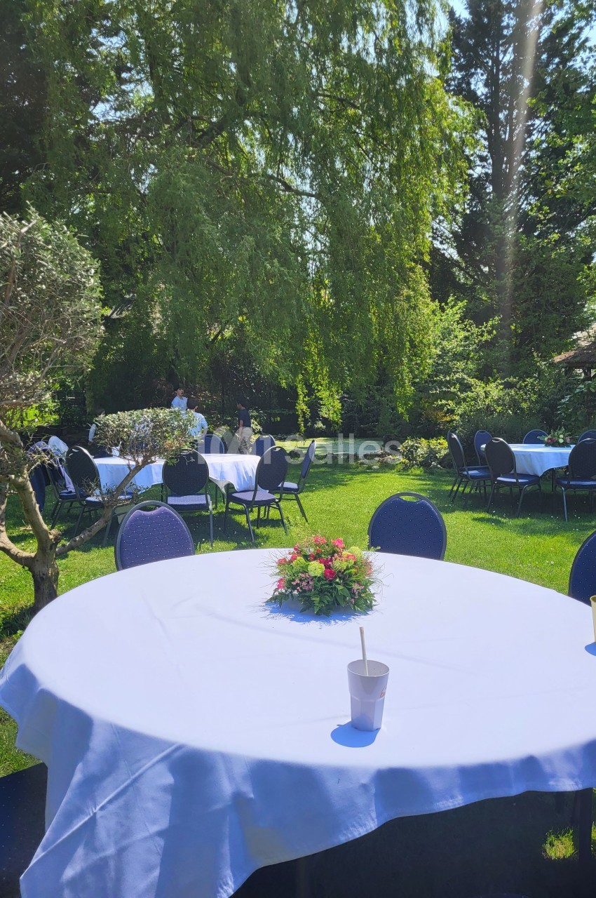 Tables rondes dressées avec nappes blanches et décorations florales dans un jardin verdoyant et ensoleillé.