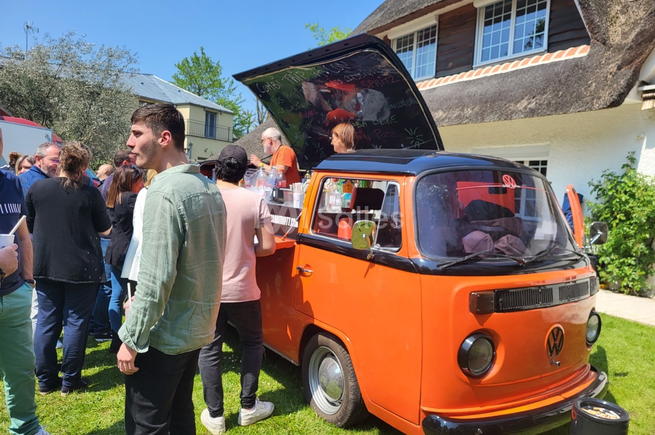 Un van orange aménagé en stand de vente, entouré de personnes dans un jardin par une journée ensoleillée.