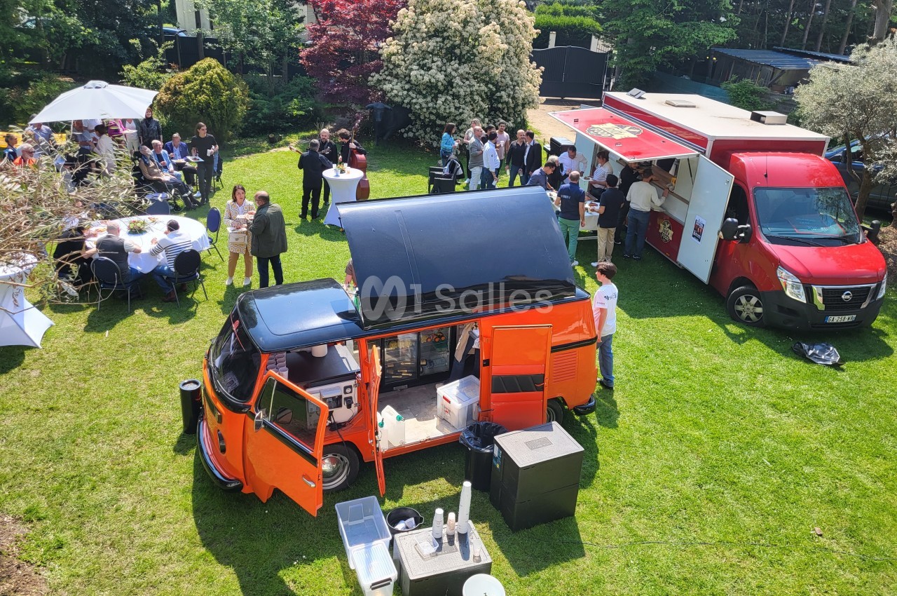 Vue aérienne d'un rassemblement en plein air avec des food trucks et des convives autour de tables sur une pelouse.