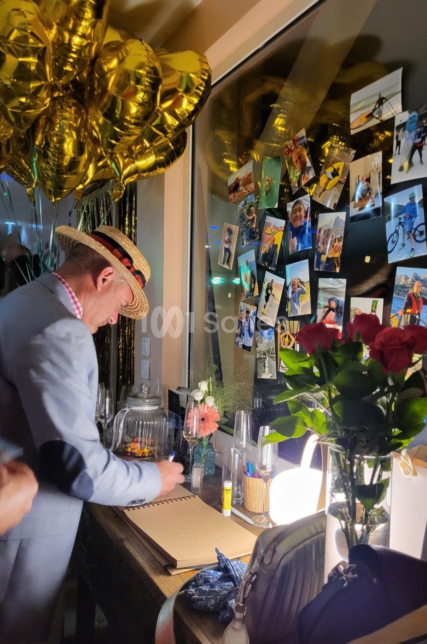 Un homme écrit dans un livre d'or lors d'un événement, entouré de ballons dorés, photos et fleurs.