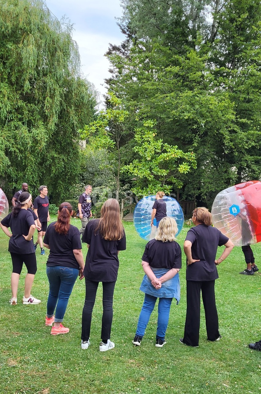 Des personnes en tenue décontractée observent un groupe jouant au bubble football dans un parc verdoyant.