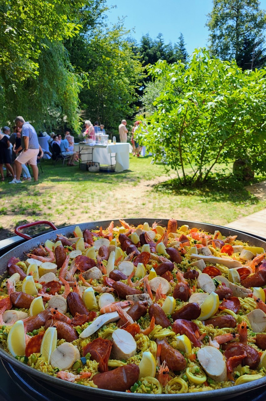 Grande poêle de paella garnie de fruits de mer, saucisses et quartiers de citron, dans un jardin avec des convives en…