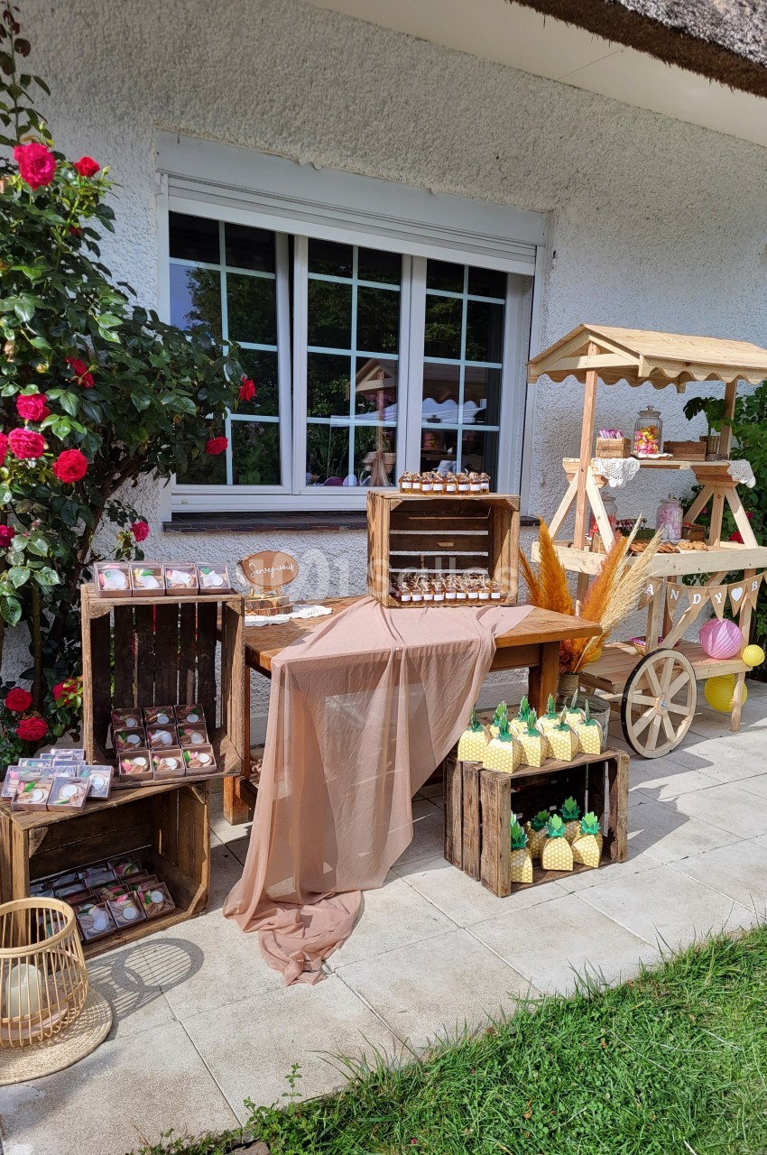 Stand extérieur décoratif avec friandises, caisses en bois, nappes en tissu et fleurs près d'une fenêtre.