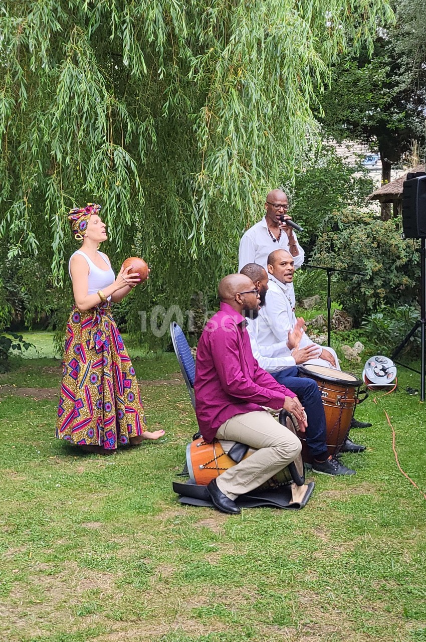Un groupe de musiciens joue des percussions en plein air, entouré de verdure et d'arbres.