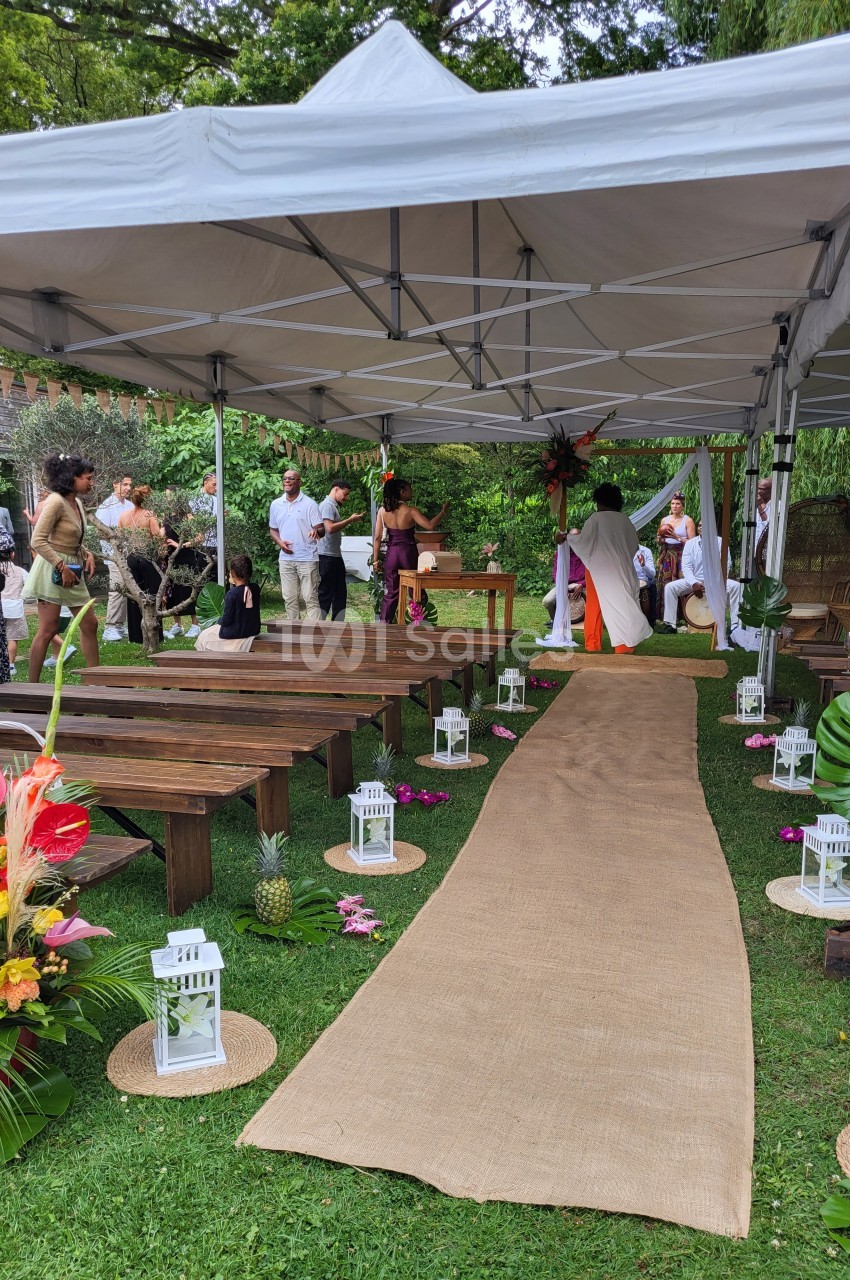 Cérémonie en extérieur sous une tente, avec allée en jute, bancs en bois et décorations tropicales.