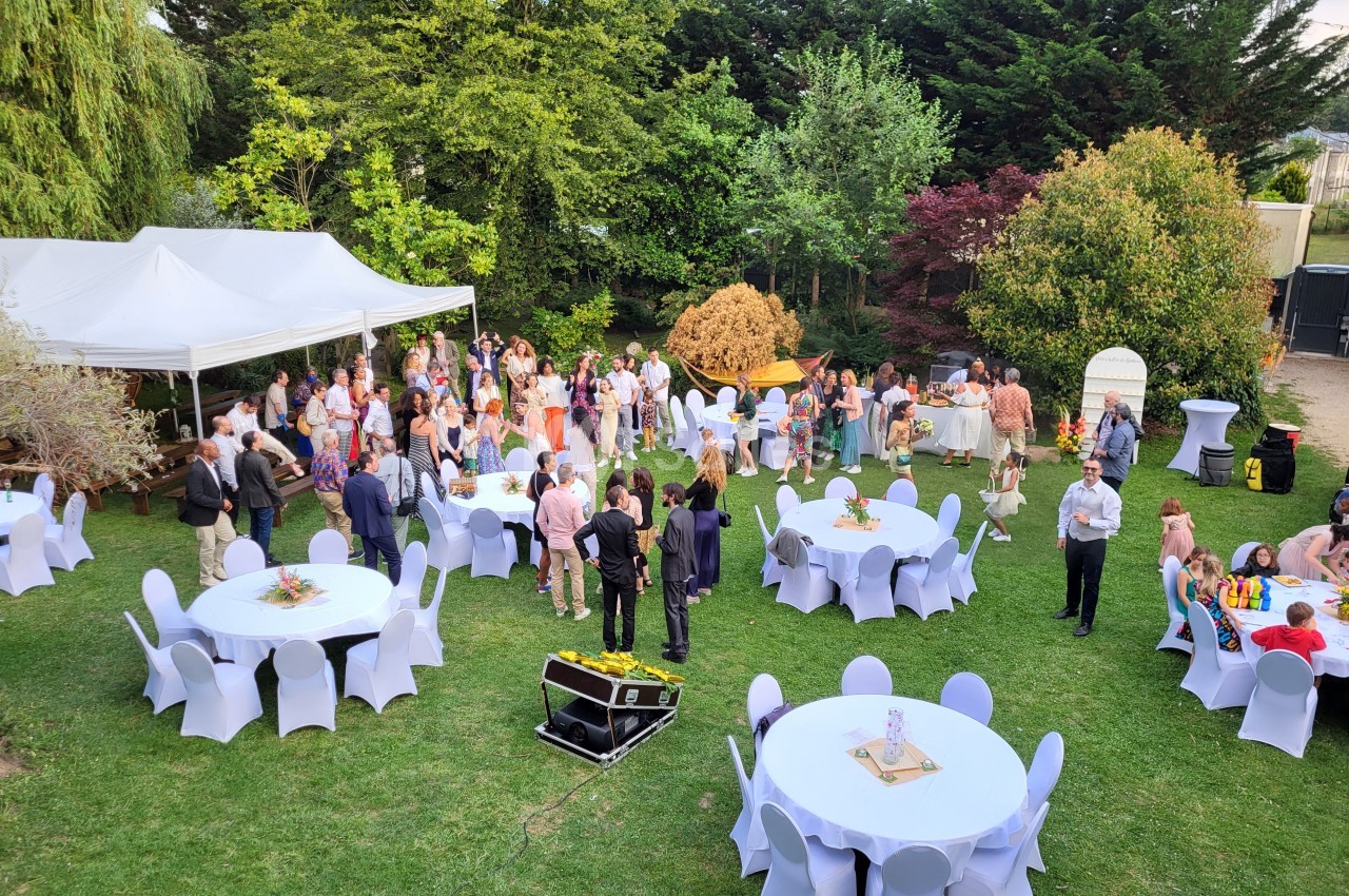 Groupe de personnes rassemblées dans un jardin avec des tables rondes dressées et une tente blanche à l'arrière-plan.