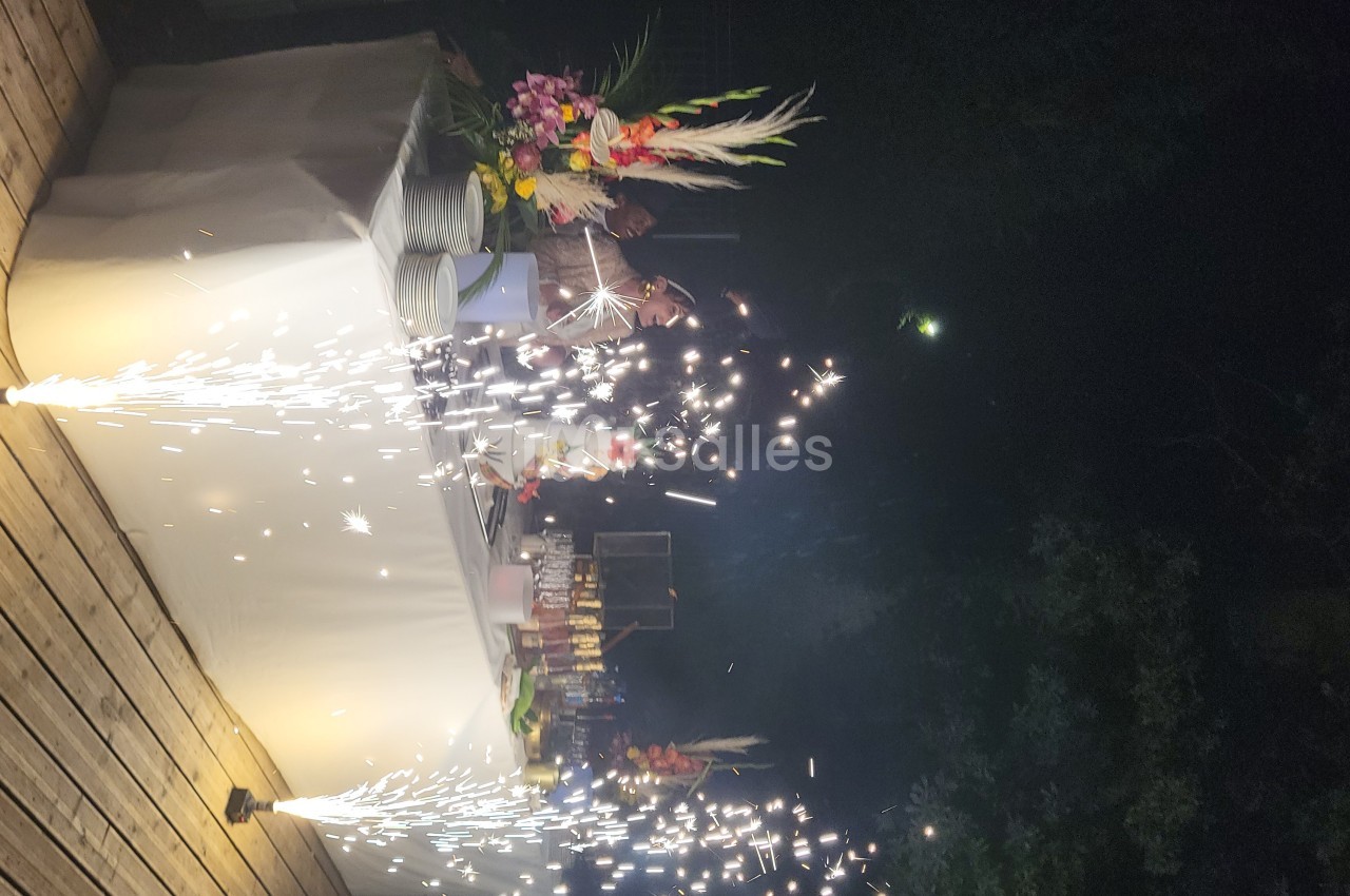 Des étincelles jaillissent devant une table décorée avec des fleurs et des plats, sur une terrasse en soirée.