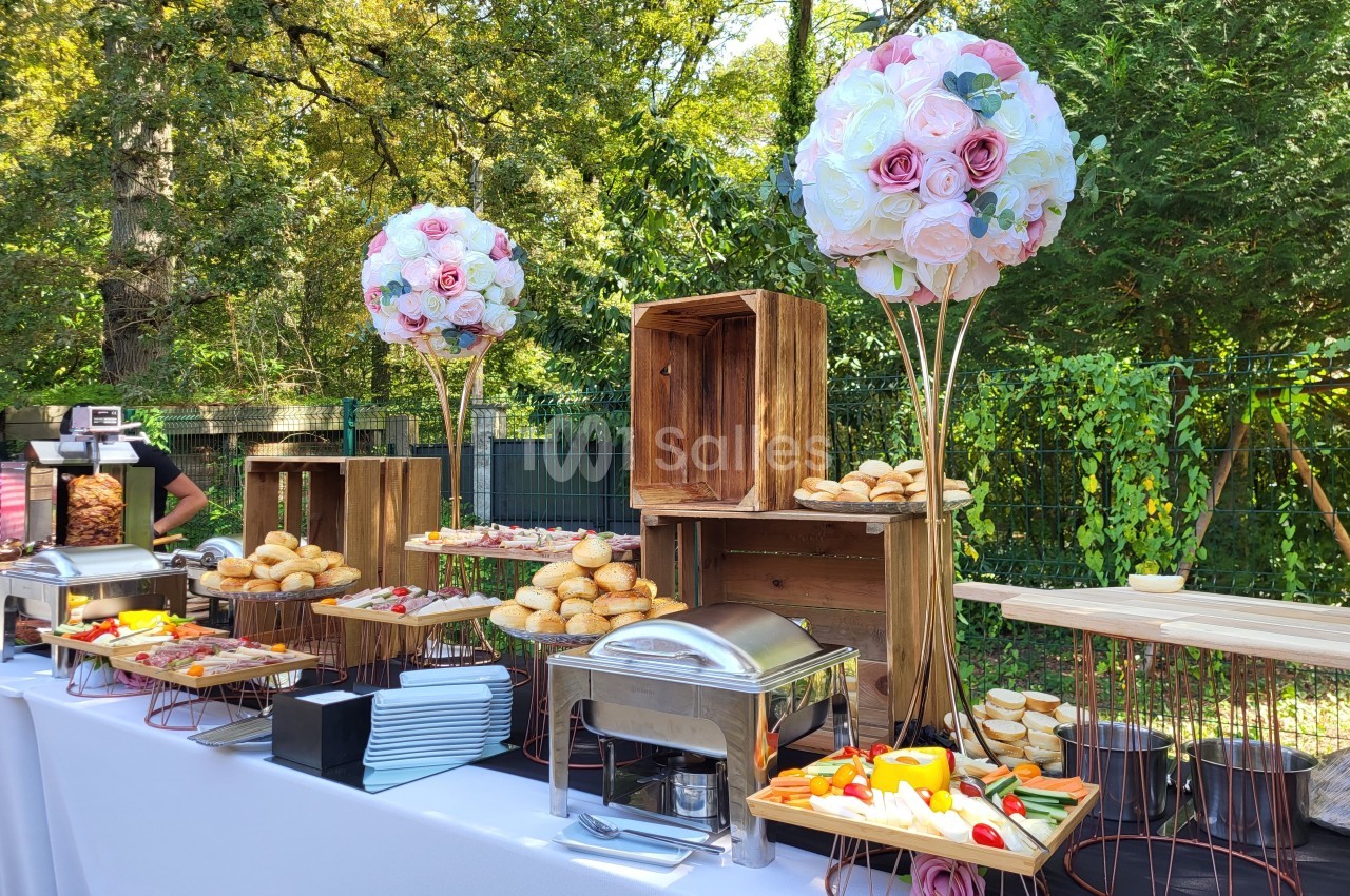 Buffet en plein air avec pains, légumes, plats chauds et décorations florales sur une table blanche.