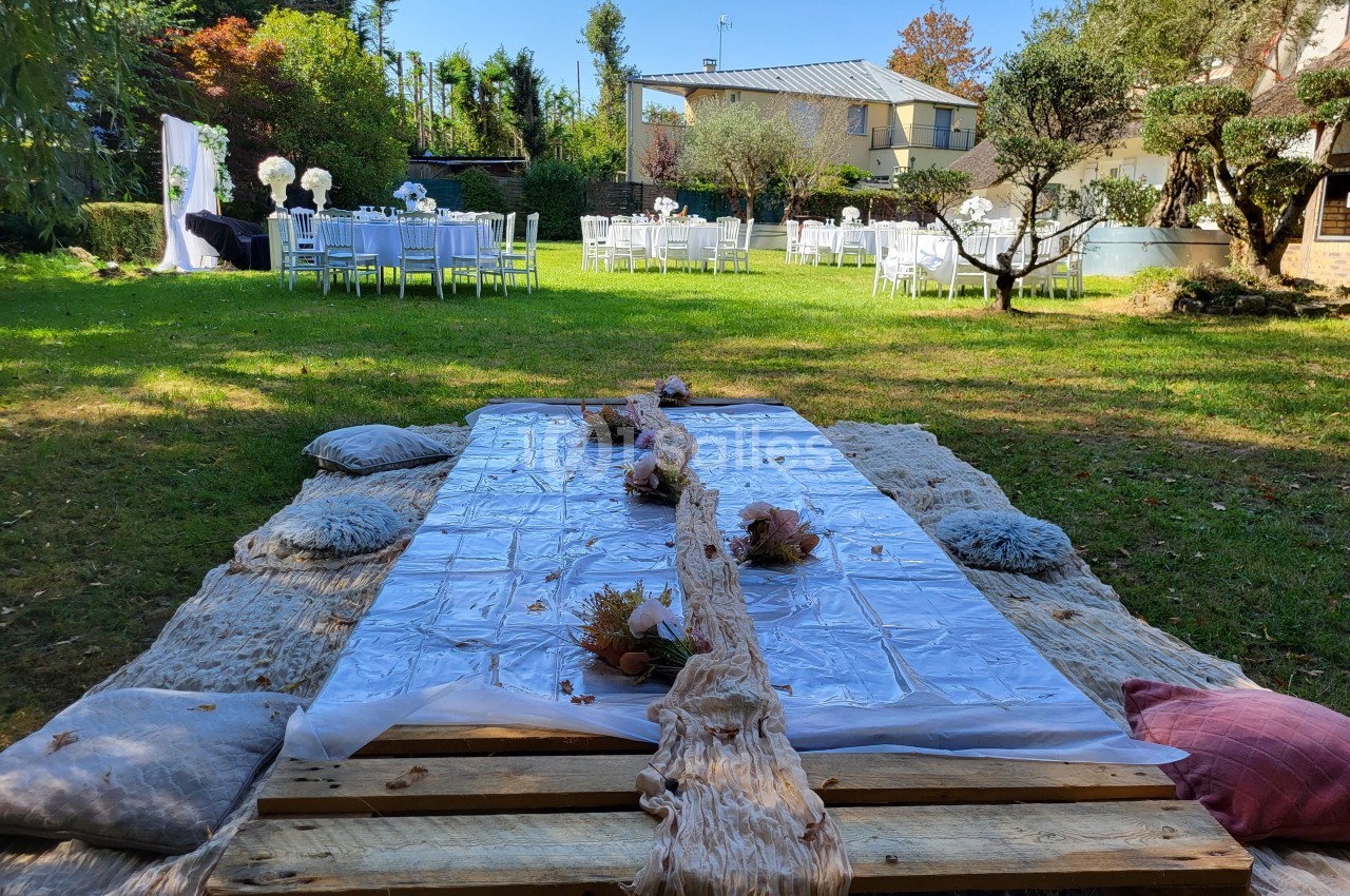 Table basse en bois décorée pour un événement en plein air, entourée de coussins, avec des tables dressées en arrière-plan.
