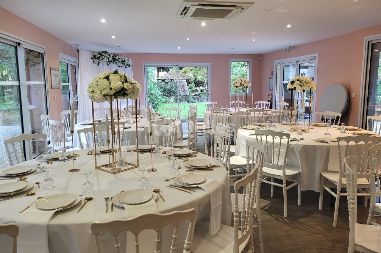 Salle de réception décorée avec des tables rondes, nappes blanches, chaises élégantes et centres de table fleuris.