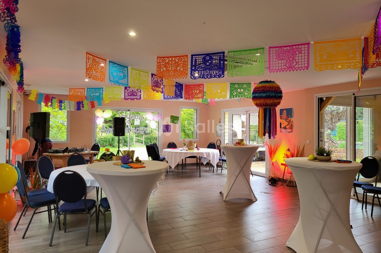 Salle décorée avec des guirlandes colorées, des tables hautes et des chaises, prête pour un événement festif.