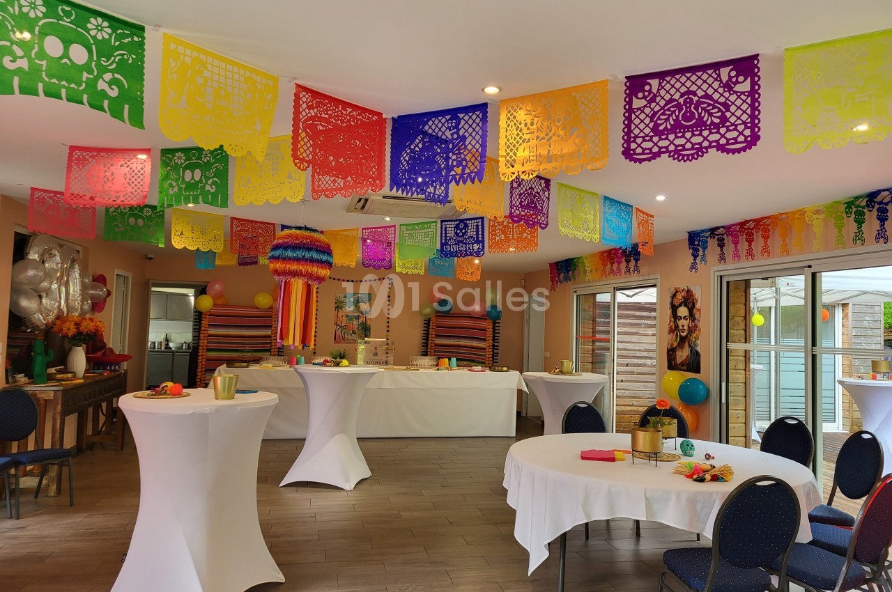 Salle décorée avec des guirlandes colorées et des tables rondes dressées pour un événement festif.