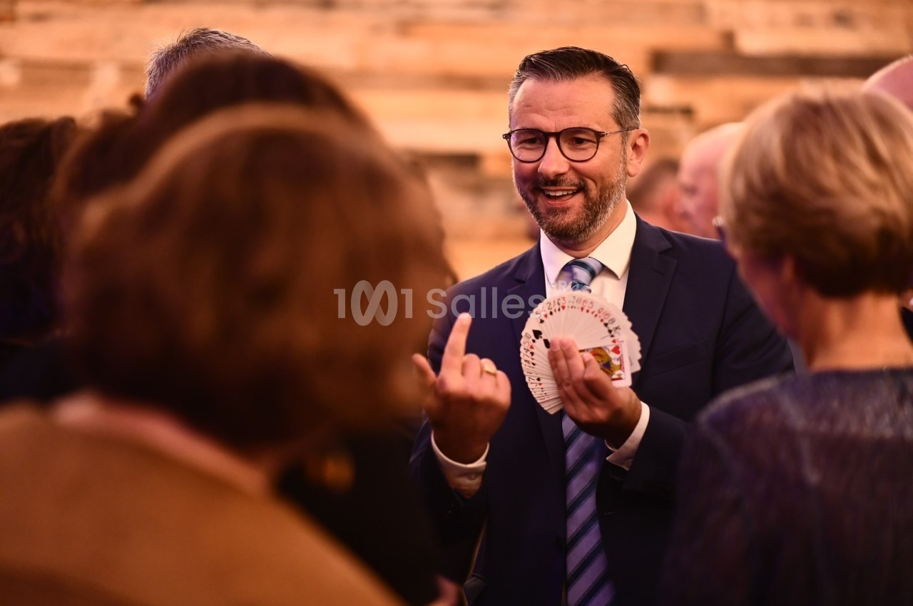 Un homme en costume réalise un tour de cartes devant un groupe de personnes dans un cadre intérieur chaleureux.