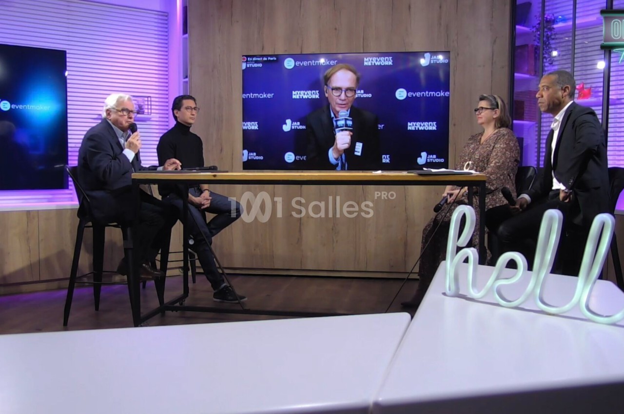 Cinq personnes discutent sur un plateau télévisé, avec un écran diffusant une intervention en visioconférence.
