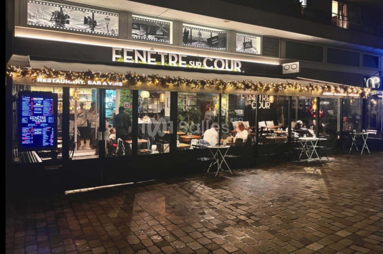 Façade d'un restaurant-bar éclairé en soirée, avec terrasse et tables extérieures sur une rue pavée.