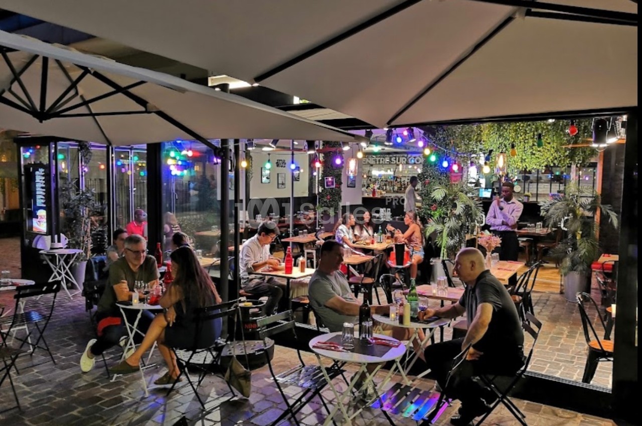 Terrasse d'un restaurant en soirée, avec des clients attablés sous des parasols et des guirlandes lumineuses colorées.