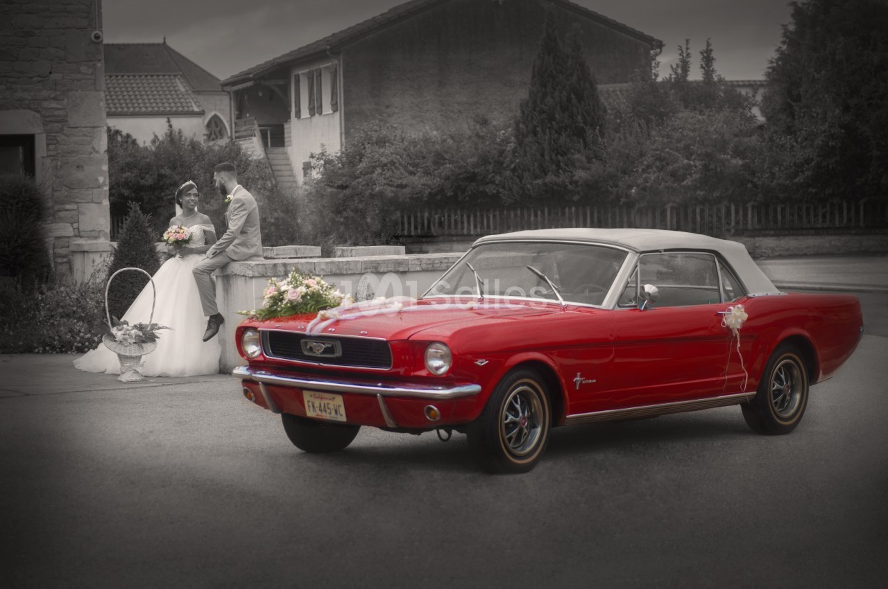 Voiture ancienne rouge avec capote blanche au premier plan, couple de mariés en arrière-plan devant un bâtiment.