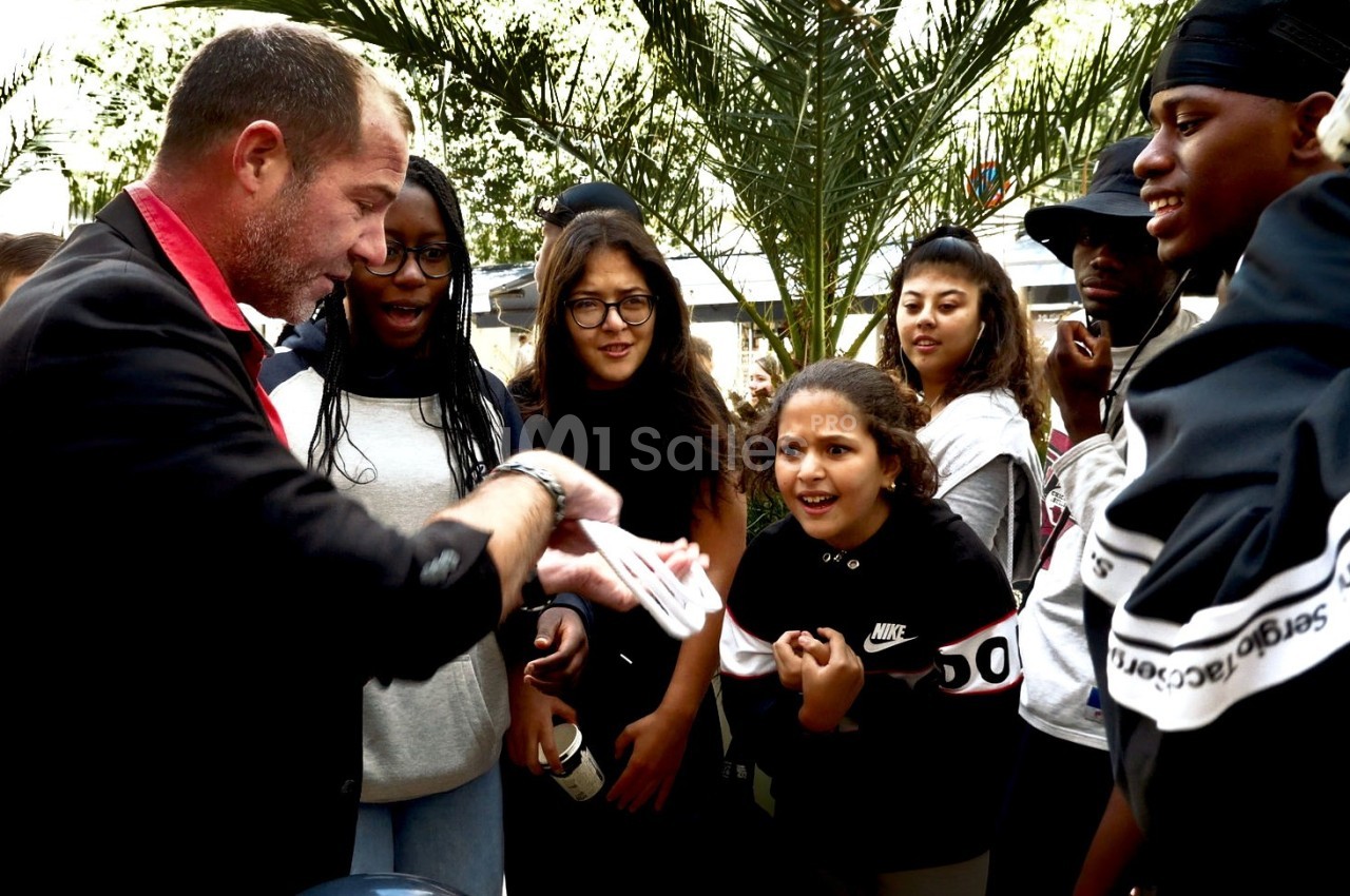 Un magicien réalise un tour de cartes devant un groupe de jeunes attentifs et surpris, en extérieur.