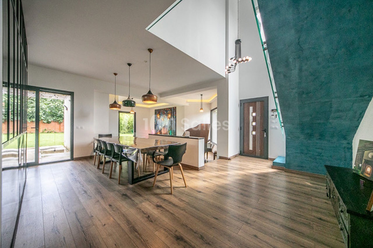 Salle à manger moderne avec table en bois, chaises noires, grandes fenêtres et escalier bleu adjacent.