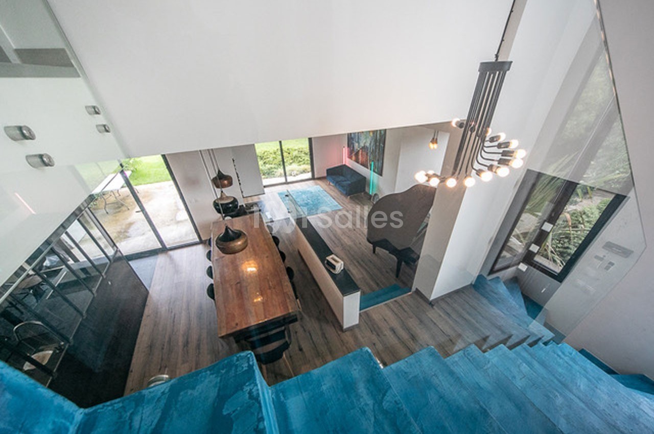 Vue plongeante sur un intérieur moderne avec salle à manger, cuisine ouverte et escalier bleu, donnant sur un jardin.