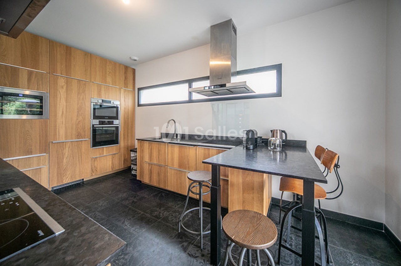 Cuisine moderne avec meubles en bois, plan de travail noir, îlot central avec tabourets et hotte en acier inoxydable.