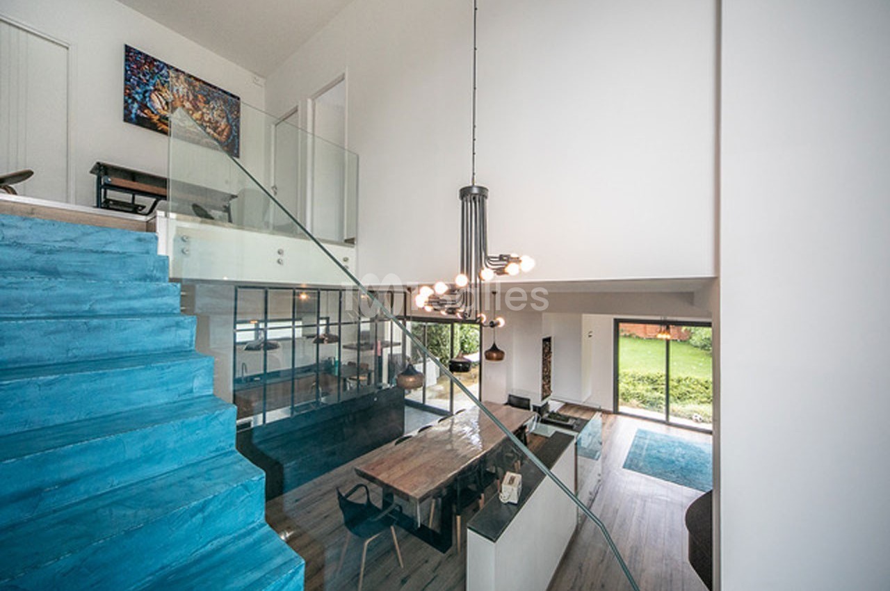 Vue intérieure d'une maison moderne avec escalier bleu, salle à manger, salon lumineux et grandes baies vitrées donnant sur…