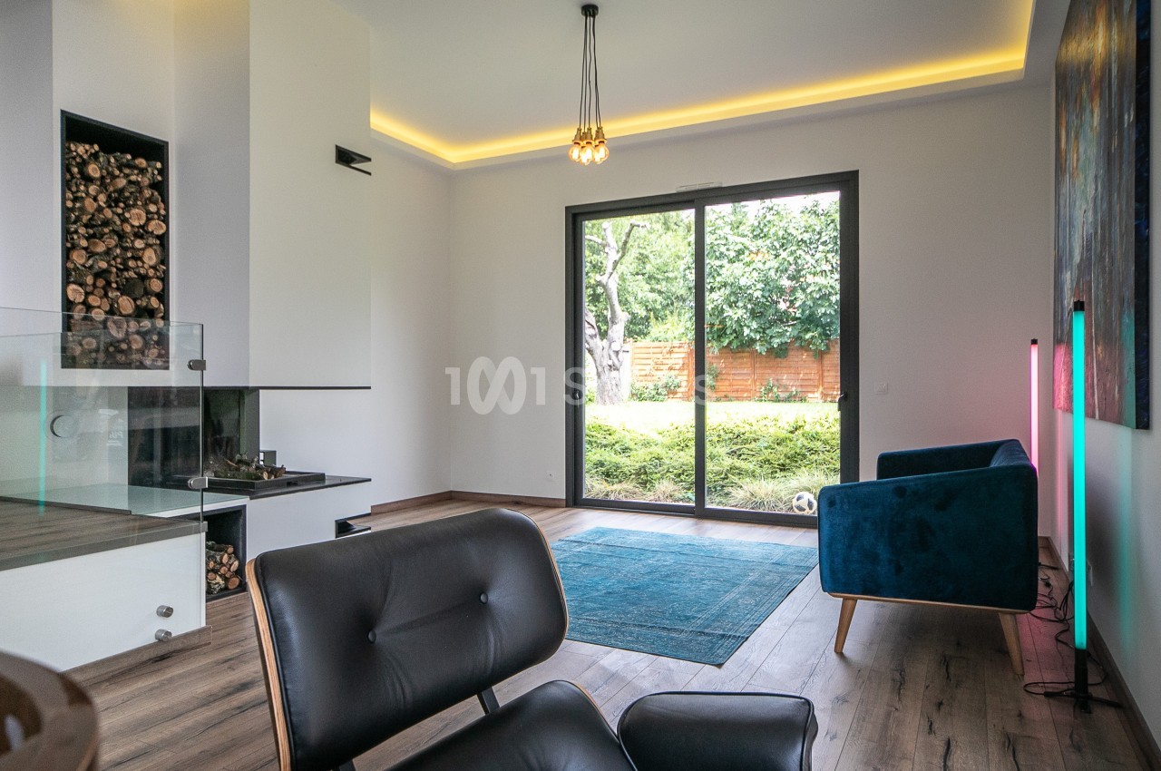 Salon moderne avec cheminée, fauteuils, tapis bleu, grande baie vitrée donnant sur un jardin verdoyant.