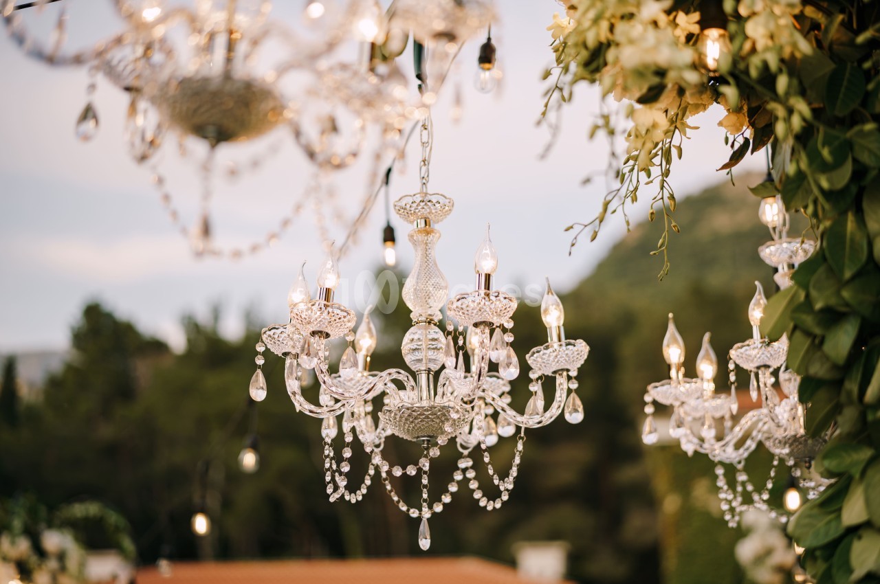 Lustre en cristal suspendu avec des ampoules allumées, entouré de verdure et d'un décor extérieur naturel.