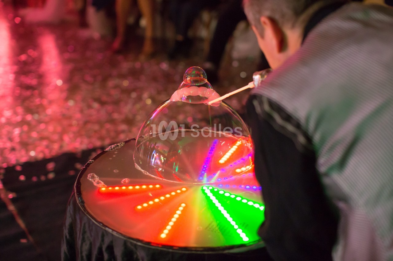 Un artiste crée des bulles de savon lumineuses sur une table éclairée par des LED colorées dans un environnement festif.